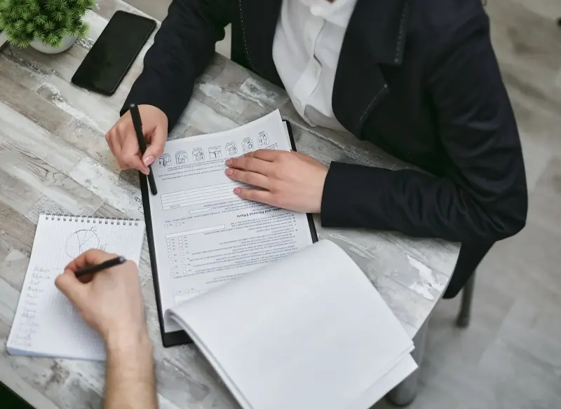 Duas pessoas revisando documentos e preenchendo formulários em uma mesa de trabalho.